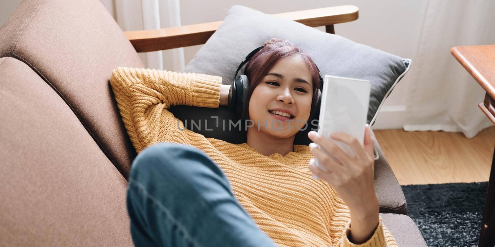 Close up smiling attractive woman in headphones using smartphone, lying on couch, positive young female looking at phone screen, watching video or making call, chatting online, listening music.