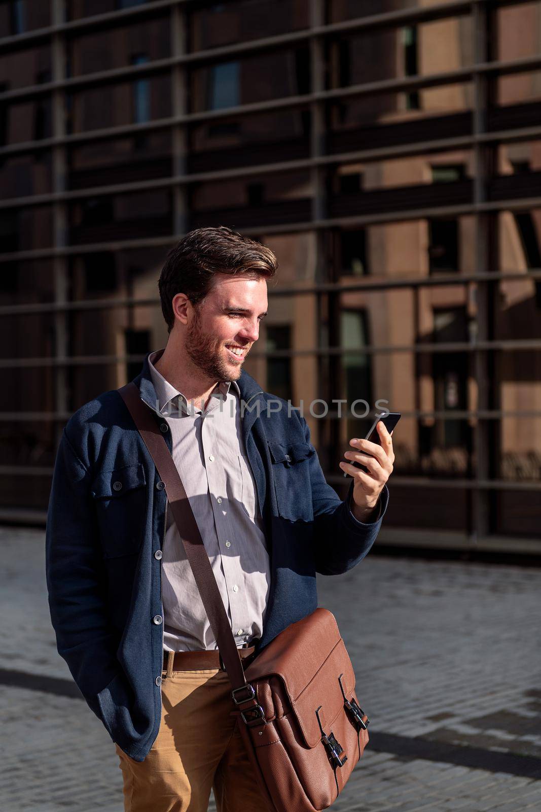 smiling young businessman looking his phone by an office building in the financial district, concept of entrepreneurship and business, copy space for text