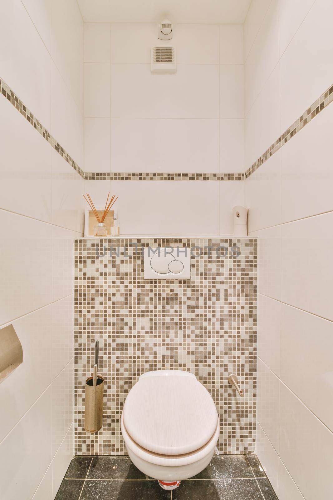 A modern toilet is installed on the wall under a button and a shelf with a diffuser in a light restroom at home