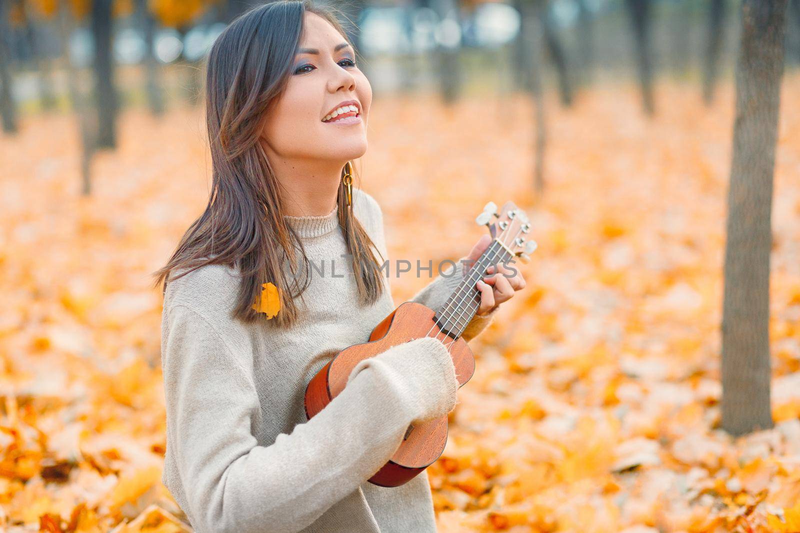 Young beautiful mixed race woman playing ukulele and singing in autumn park.