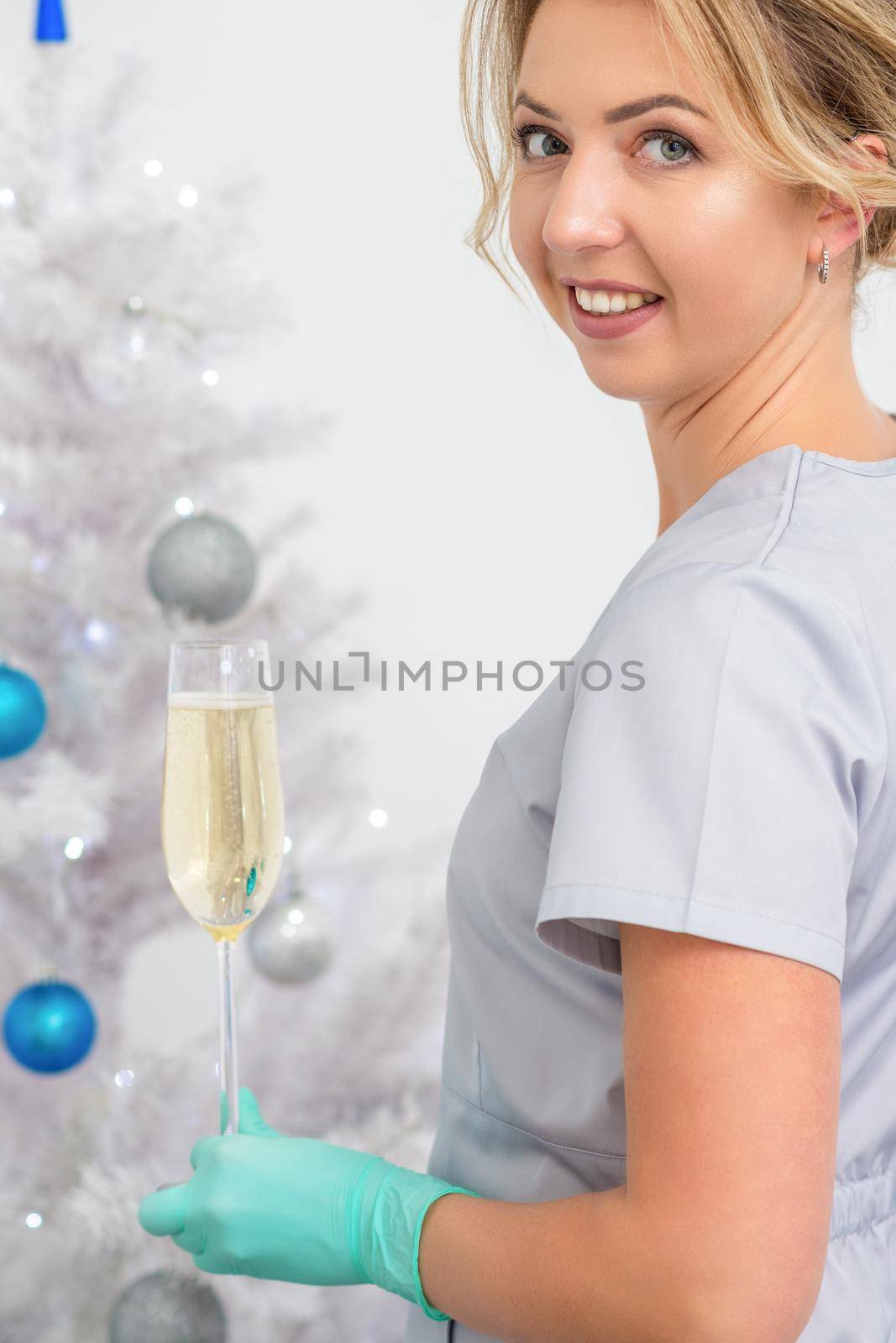 Portrait of female doctor with glass of champagne standing near the christmas tree looking at camera