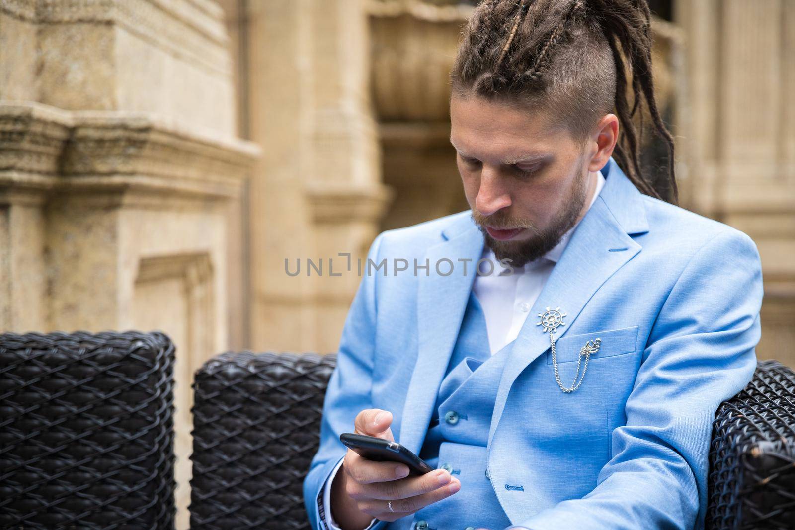 A young hipster man with dreadlocks in a blue suit sits in a street restaurant looking at a smartphone, sad and upset.