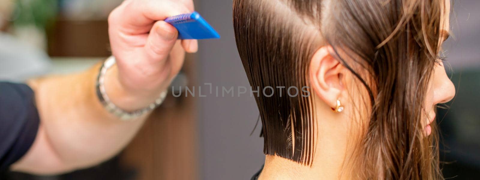 Side view of hand of hairdresser doing haircut of young woman by comb in hair salon. Selective focus