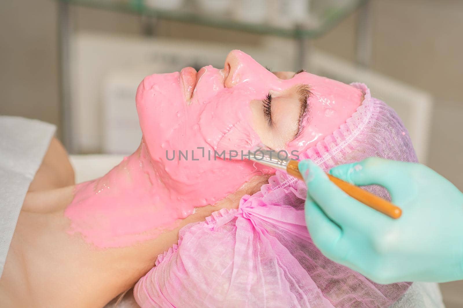 Hand of cosmetologist applying pink alginic mask to face of young woman in beauty salon