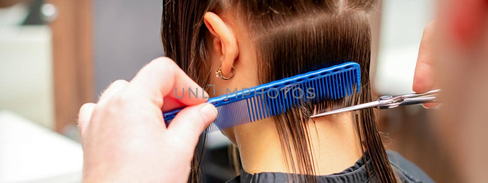 Back view of hands of male hairdresser cuts off long hair of young woman in hair salon. Selective focus