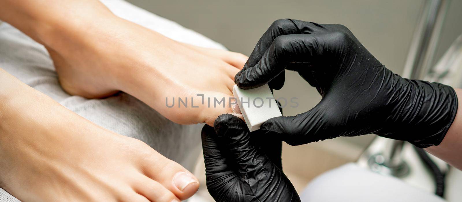 Hands of pedicurist buffing toenail of woman by white nail buff in nail salon