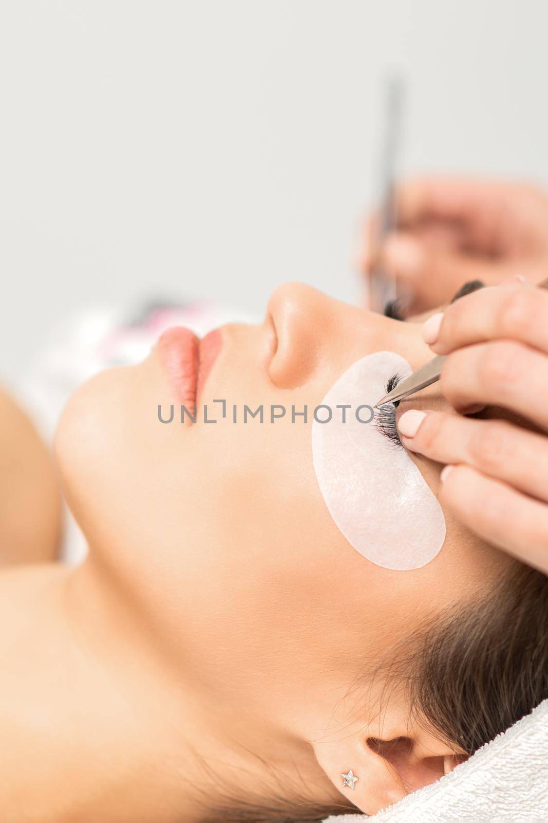 Closeup view of young caucasian woman having eyelash extension procedure in beauty salon. Beautician glues eyelashes with tweezers