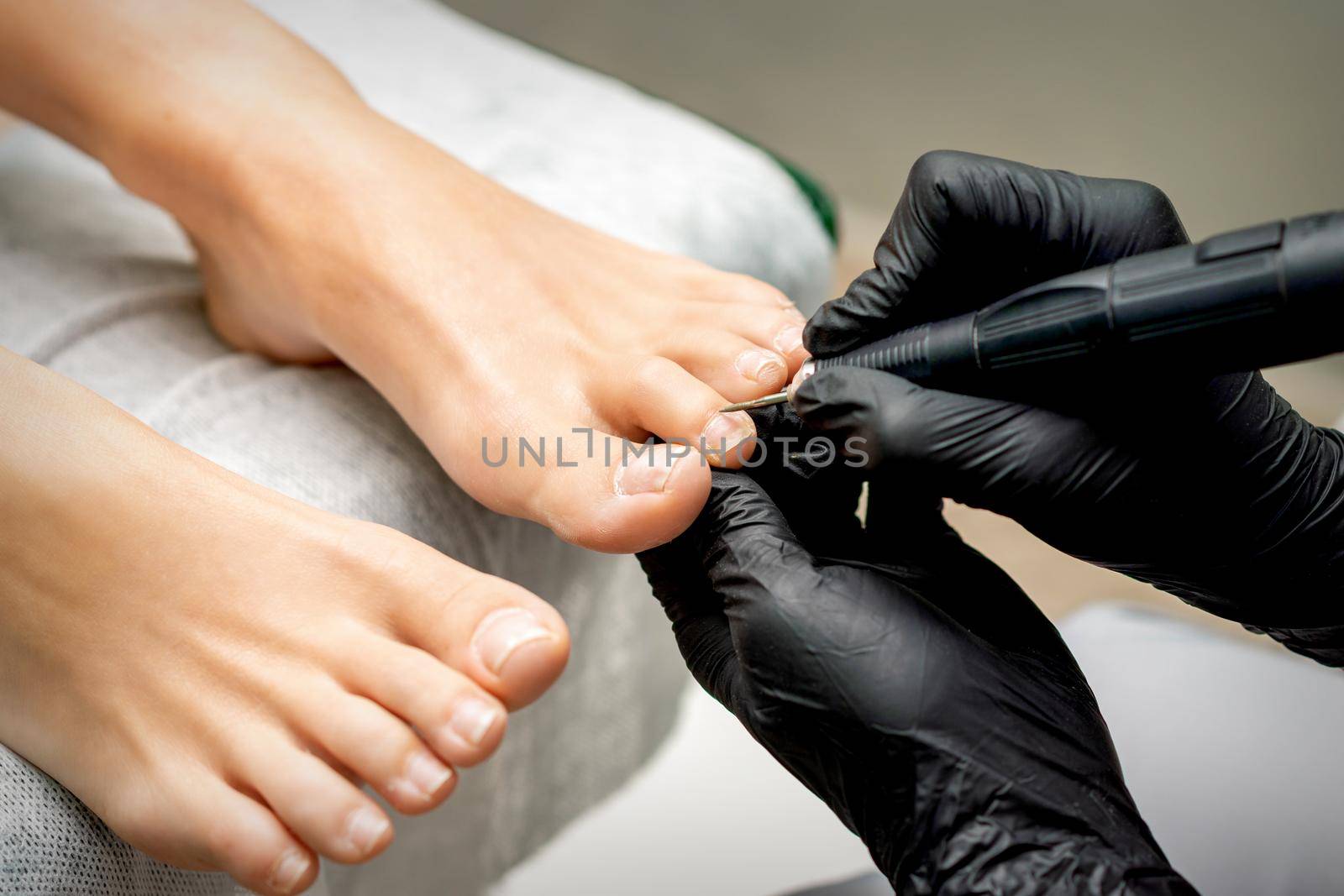 Pedicure master removes cuticle from toes of woman using professional electric nail hardware in nail salon