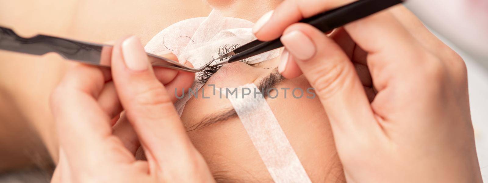 Young caucasian woman having eyelash extension procedure in beauty salon. Beautician glues eyelashes with tweezers