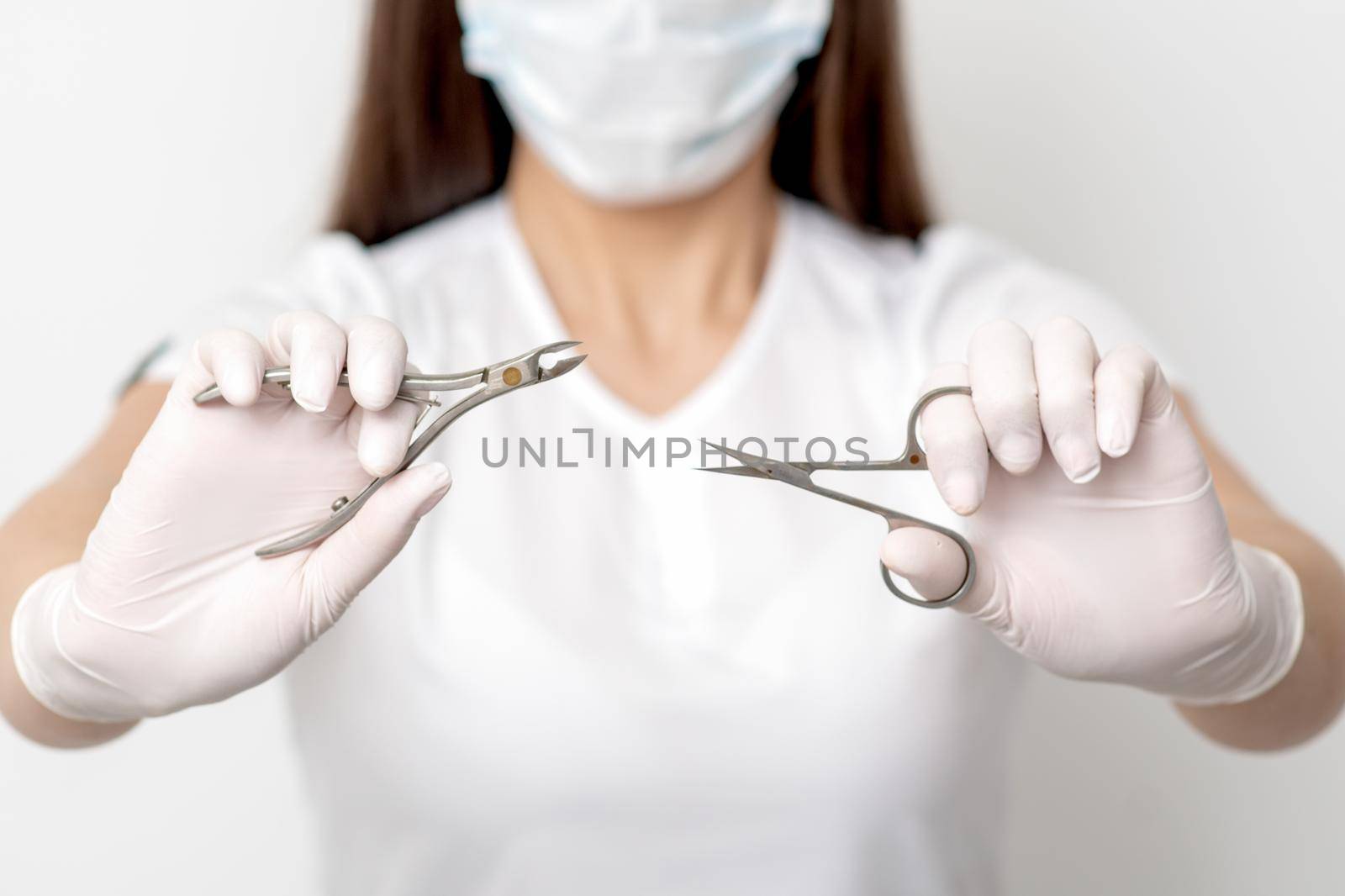 Manicure tools in hands of female manicurist wearing white mask and gloves on white background