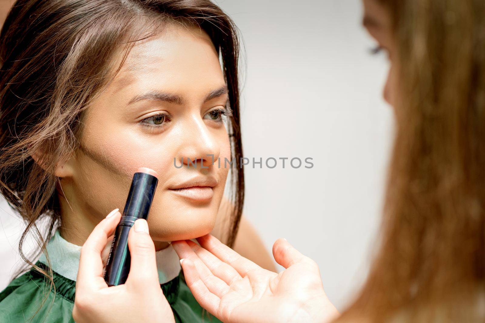 Makeup artist applying cream blush stick foundation on cheek of young caucasian woman