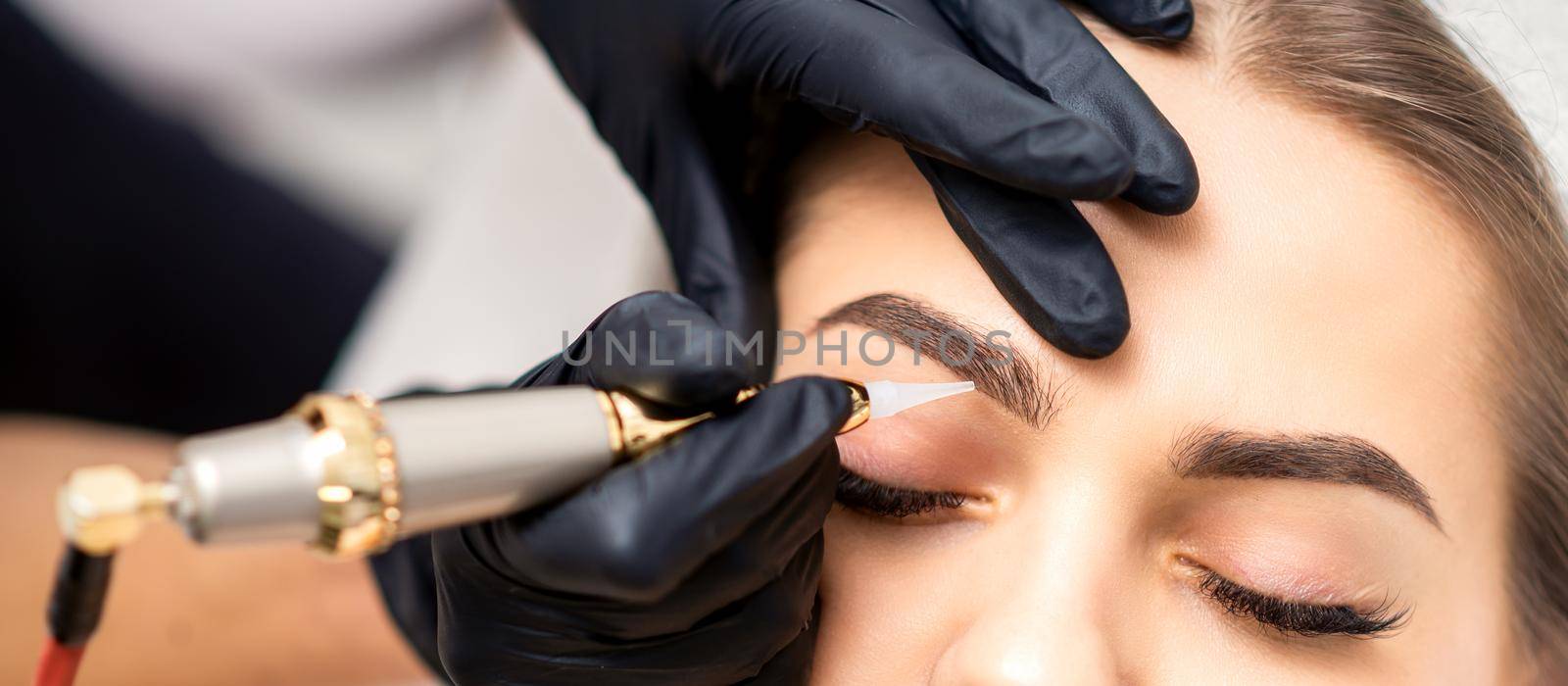 Beautician applying permanent makeup on eyebrows of young woman by special tattoo machine tool