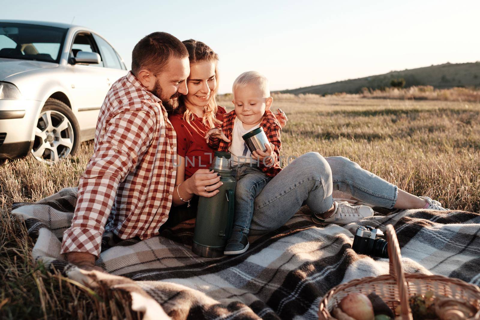 Happy Young Family Mom and Dad with Their Little Son Enjoying Summer Weekend Picnic Sitting on the Plaid Near the Car Outside the City in Field at Sunny Day Sunset, Vacation and Road Trip Concept