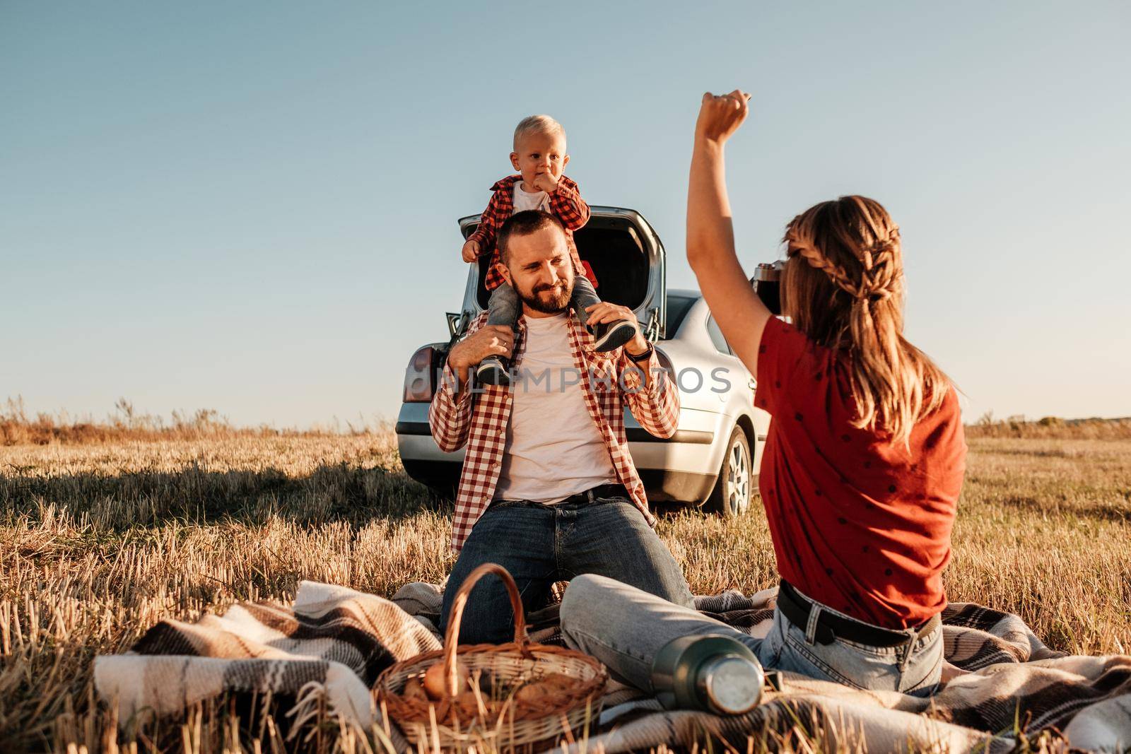 Happy Young Family Mom and Dad with Their Little Son Enjoying Summer Weekend Picnic Sitting on the Plaid Near the Car Outside the City in Field at Sunny Day Sunset, Vacation and Road Trip Concept