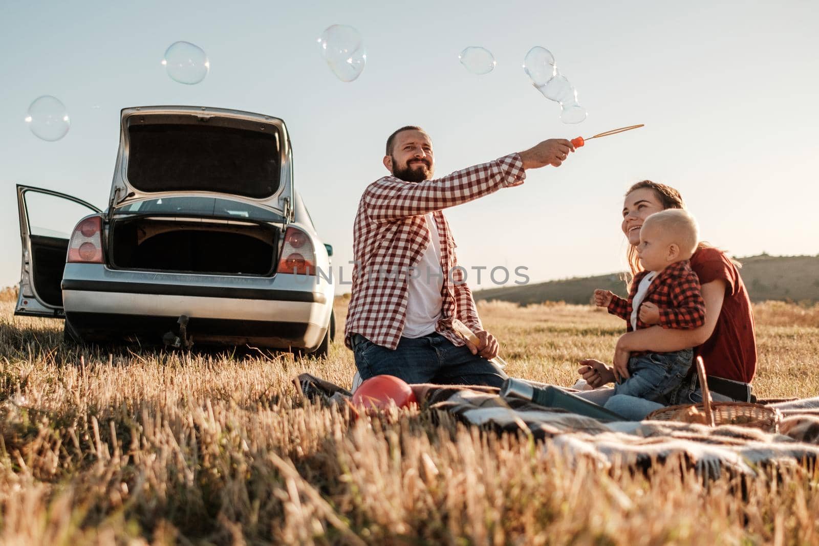 Happy Young Family Mom and Dad with Their Little Son Enjoying Summer Weekend Picnic on Car Outside the City, Playing with Bubbles