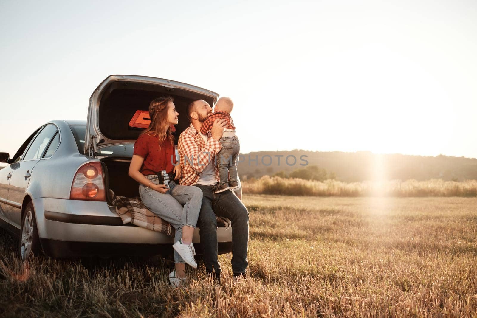 Happy Young Family Mom and Dad with Their Little Son Enjoying Summer Weekend Picnic Sitting on the Trunk of the Car Outside the City in Field at Sunny Day Sunset, Vacation and Road Trip Concept
