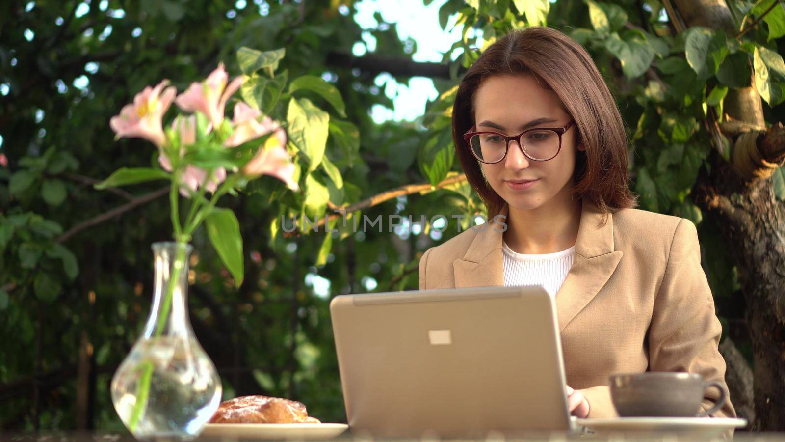 Young business woman sits in a cafe and works at a laptop. Girl with coffee and a bun on the summer veranda. 4k