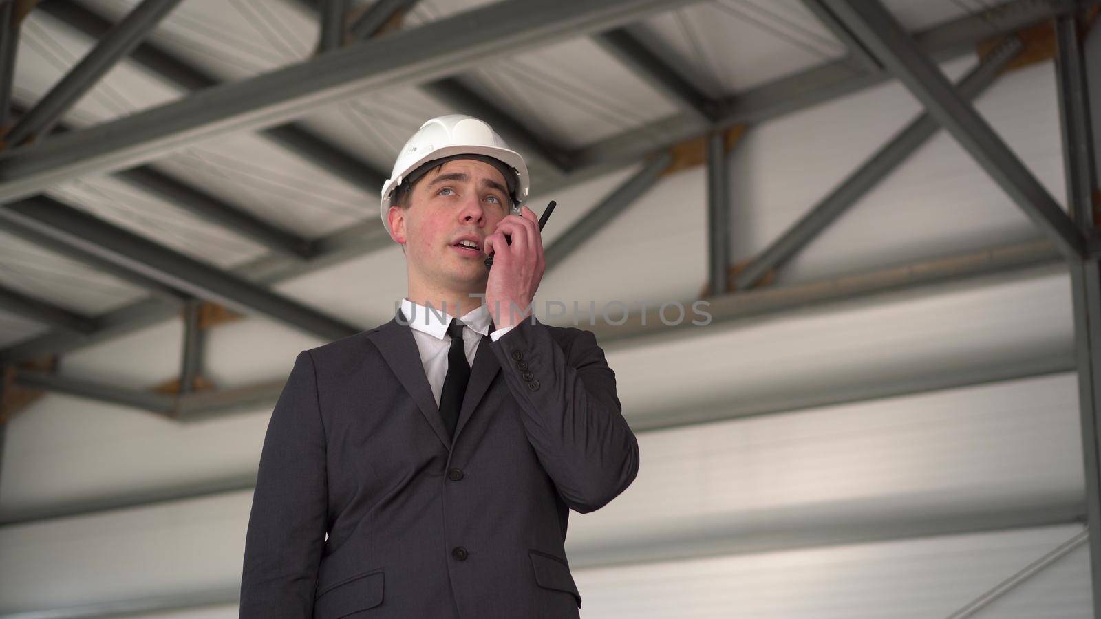 A young man in a helmet speaks on a walkie-talkie at a construction site. The boss in the suit looks around. 4k