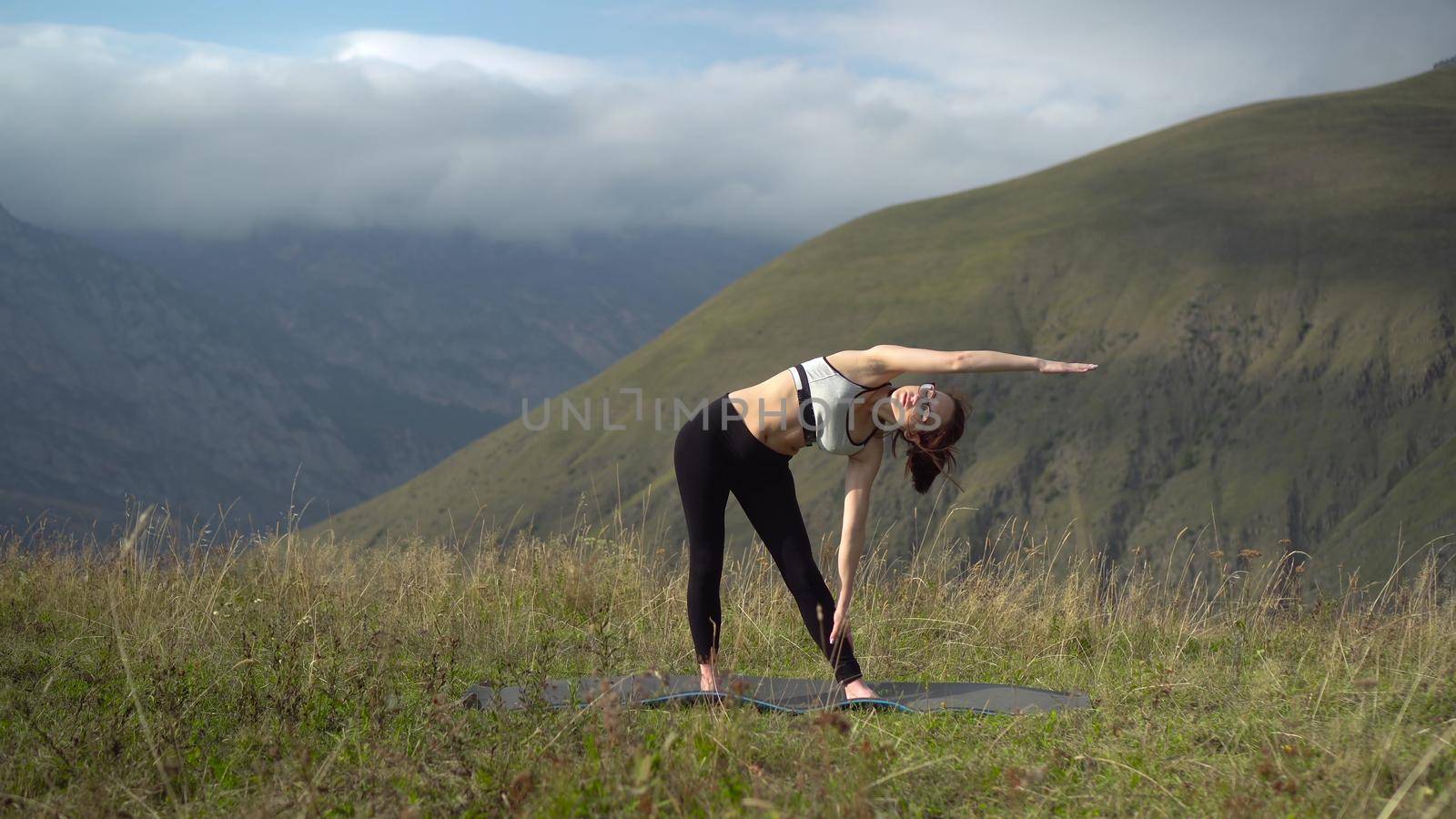Young woman in tracksuit is engaged in yoga performing triconasana pose in the mountains. The camera moves to create a parallax effect. by Puzankov