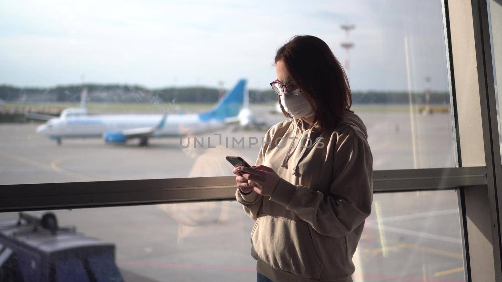 Young woman in a medical mask with a phone in her hands on the background of a window at the airport. Airplanes in the background. by Puzankov