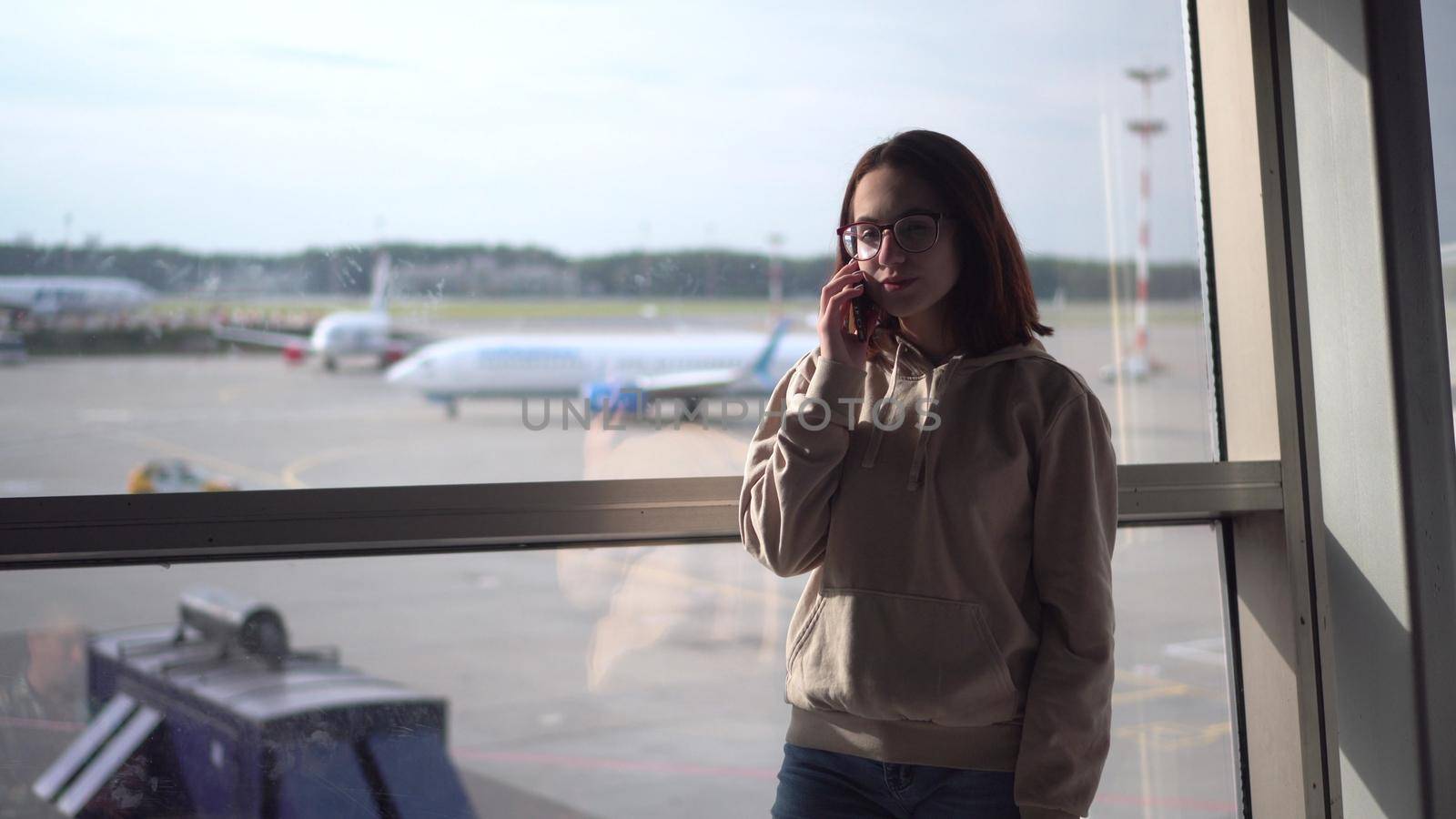 A young woman in a sweatshirt and glasses speaks on the phone against the background of a window at the airport. Airplanes in the background. by Puzankov