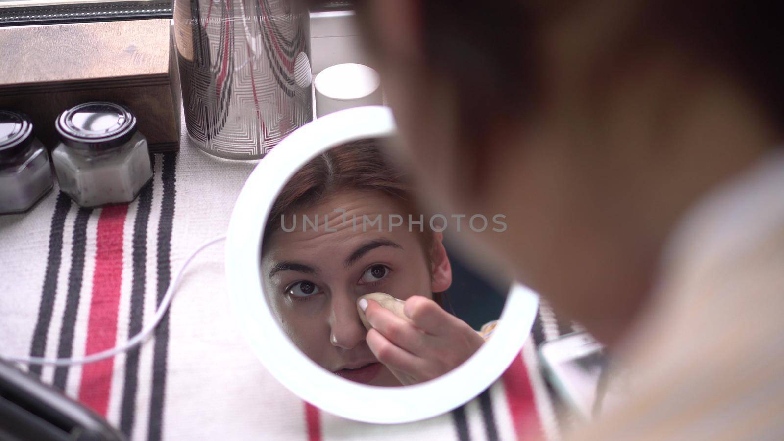 A young woman in front of a window looks into a small mirror with light and applies makeup with a sponge. View of the girl through the mirror. 4k