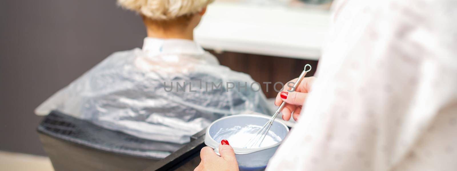 Hairdresser is preparing white dye in container on female client background