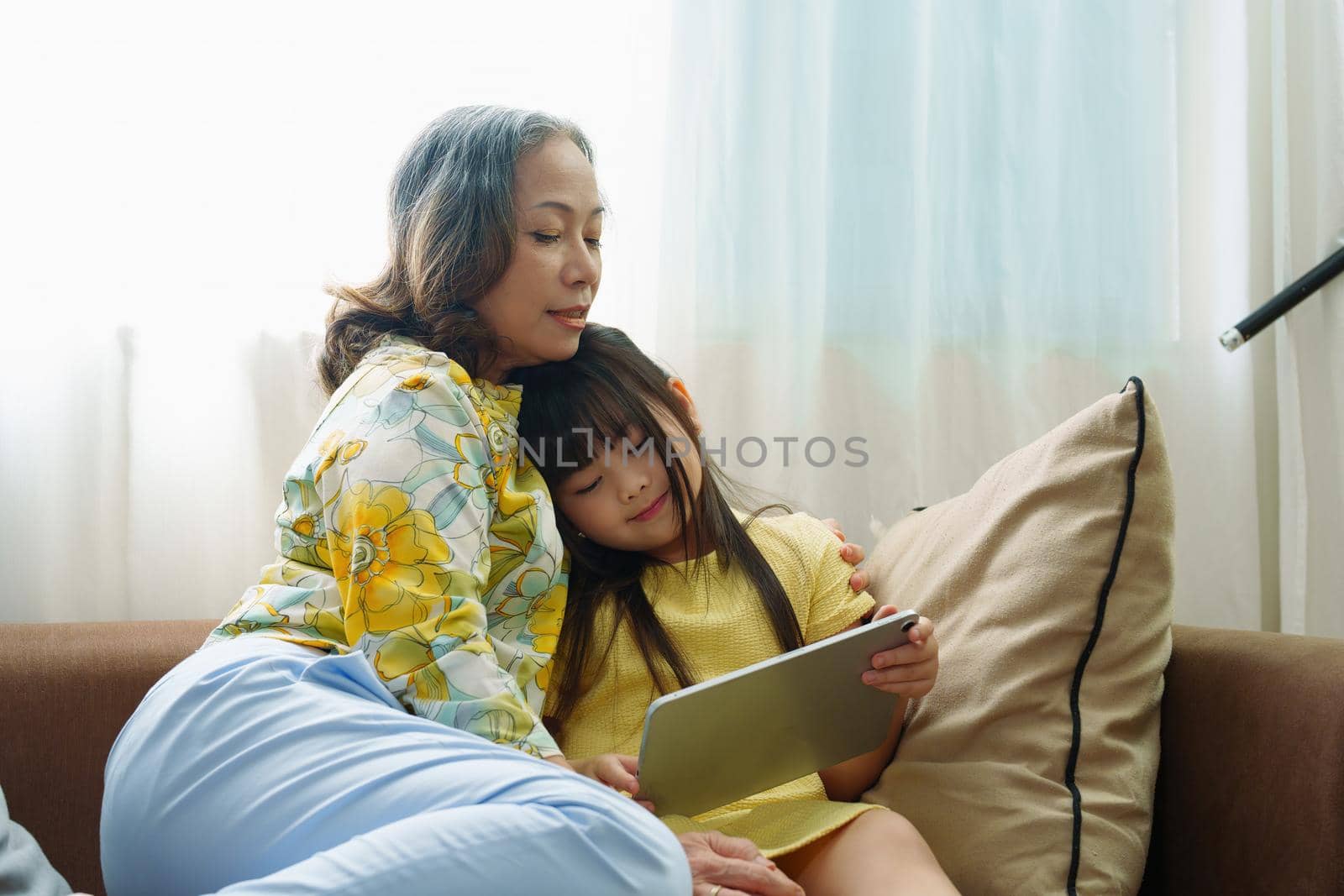 Asian portrait, Grandma and granddaughter doing recreational activities playing tablet computers on the sofa by Manastrong