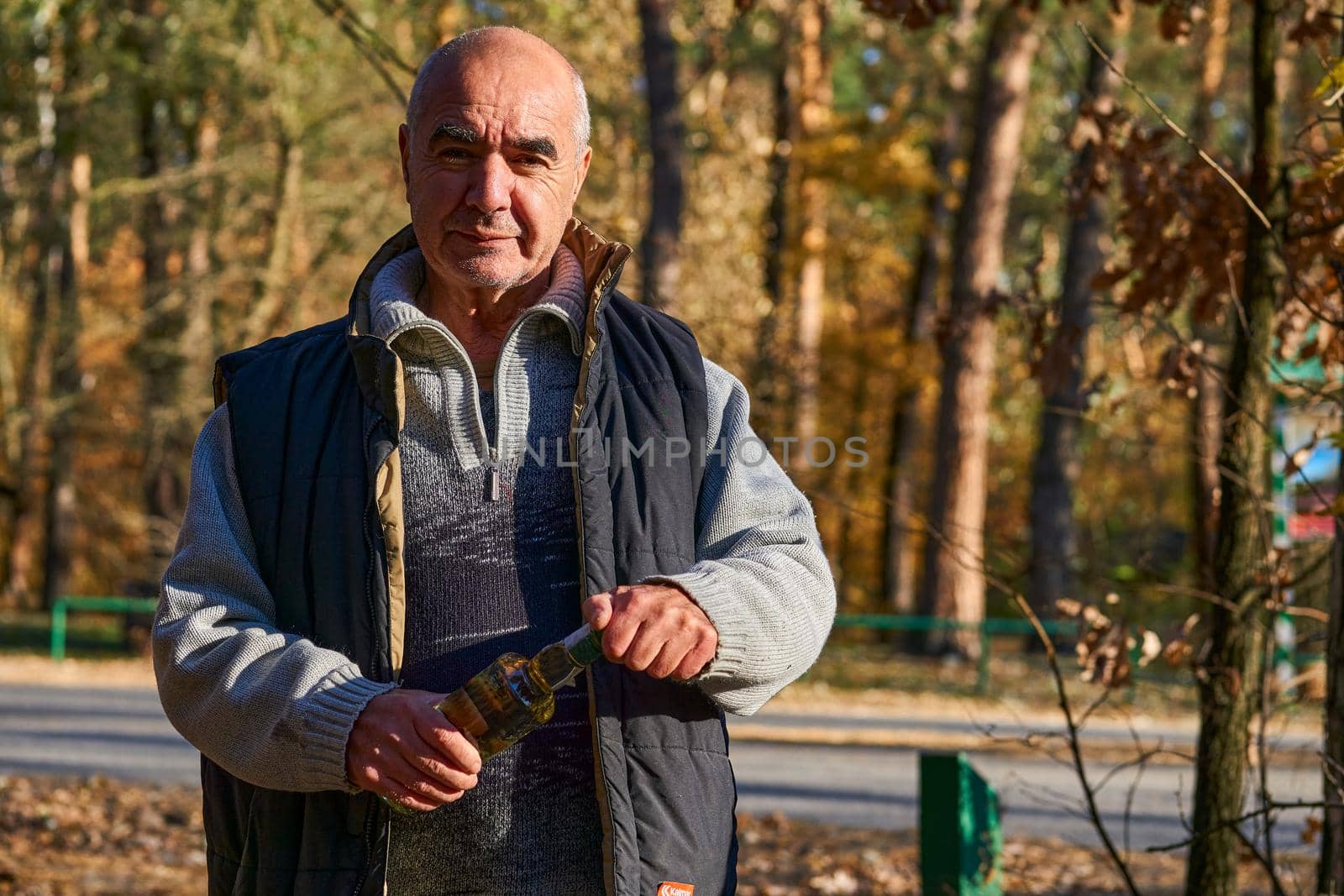 Cheerful elderly male pensioner with a bottle of alcohol in nature on sunny day by jovani68