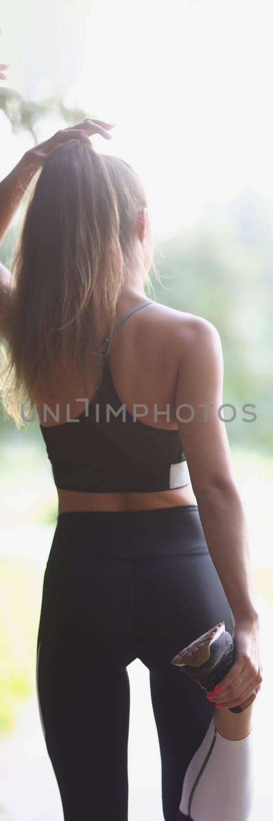 Close-up of young woman stand with back and stretching leg pulling it with hand. Active sportswoman in outfit for sport activity. Active lifestyle concept