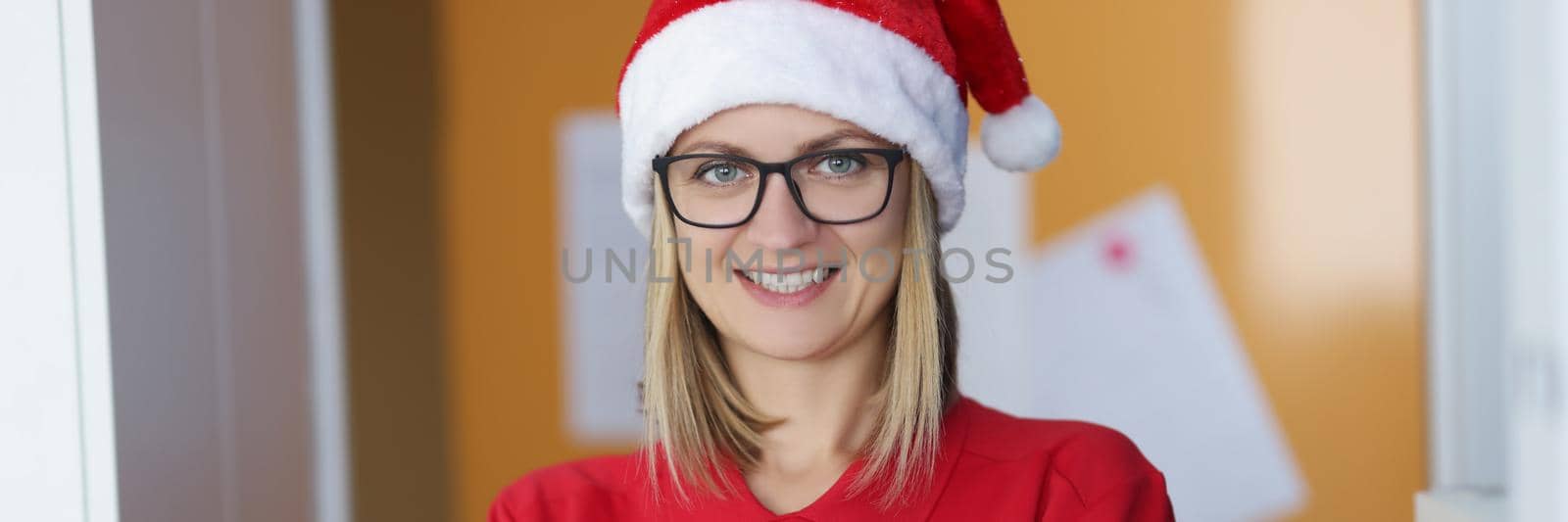 Portrait of happy blonde lady courier in red shirt and santa claus hat. Positive mood, festive outfit to meet new year. Delivery service, christmas concept