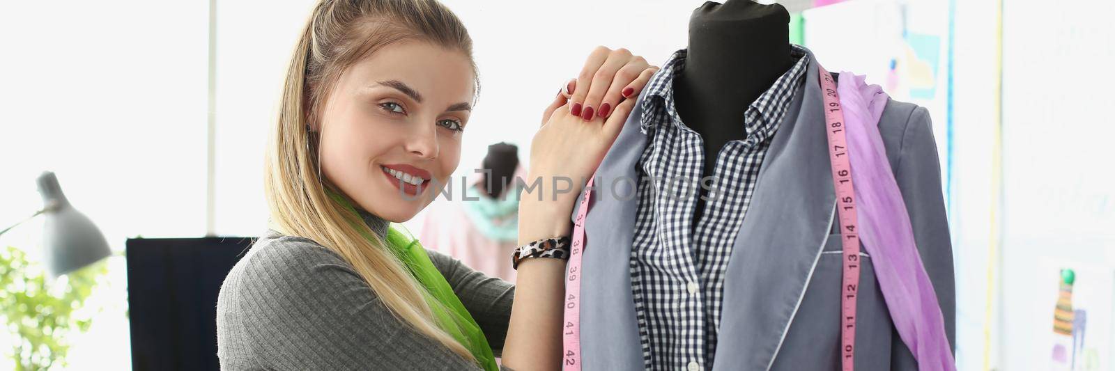 Portrait of woman seamstress posing near brand new jacket and being proud of it. Smiling designer owns successful atelier. Sew, tailoring, fashion concept