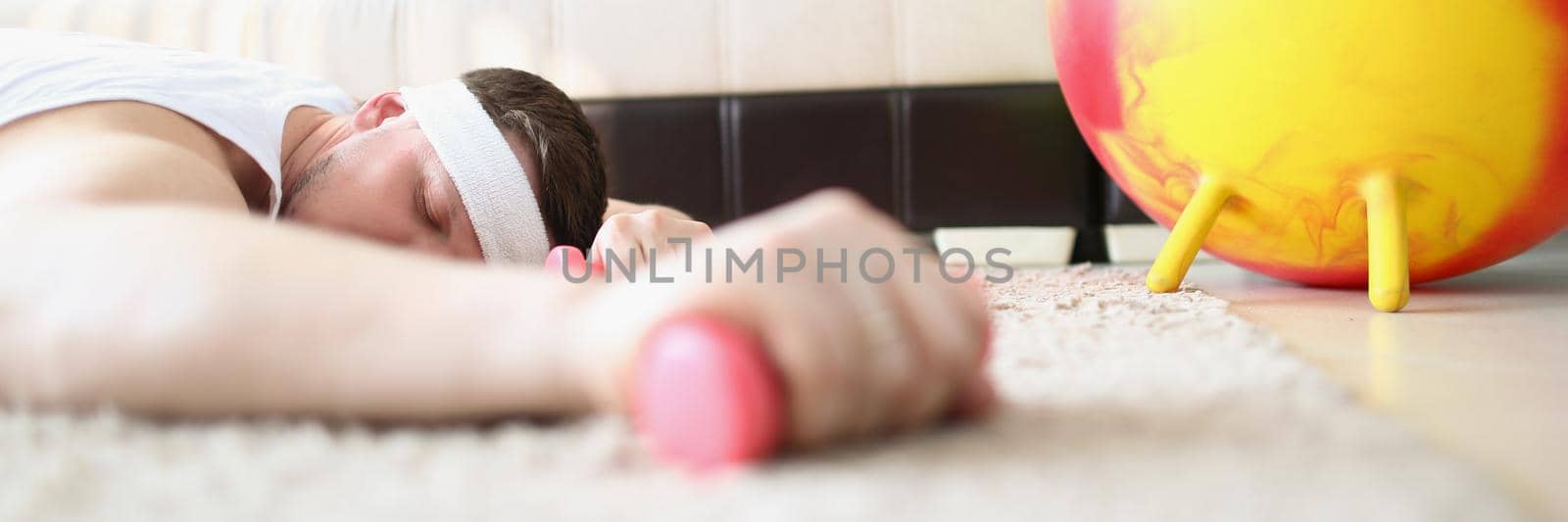 Close-up of tired man laying on carpet floor with dumbbell equipment in hand. Exhausted sportsman after hard exercises fell asleep. Home, sport concept