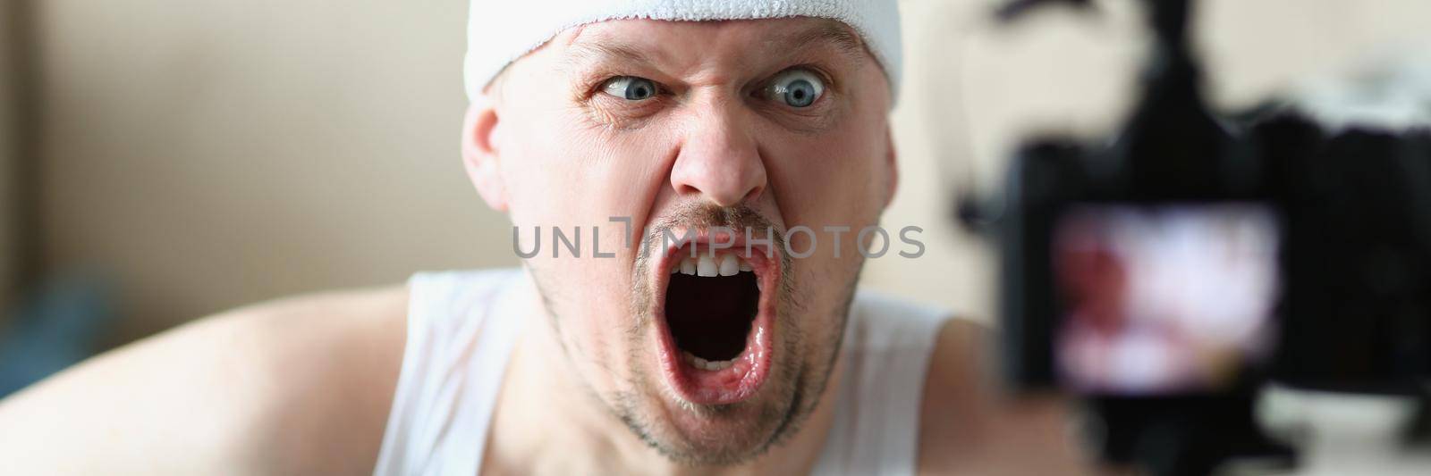 Portrait of man screaming on camera while performing exercise at home. Man in sportswear training indoors, film class on video. Sport, work hard concept