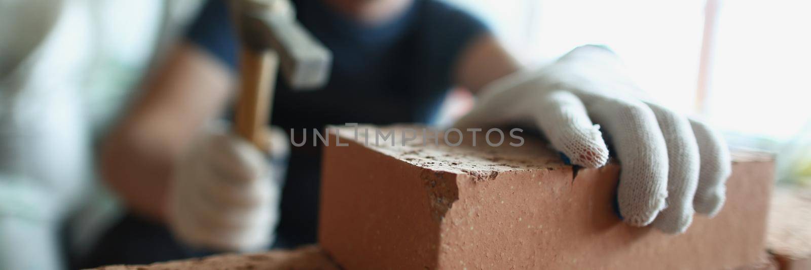 Close-up of skilled foreman in hardhat use hammer equipment to put blocks on unfinished structure. Big concrete wall made of red bricks. Renovation concept
