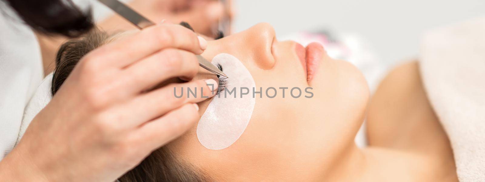 Young caucasian woman having eyelash extension procedure in beauty salon. Beautician glues eyelashes with tweezers