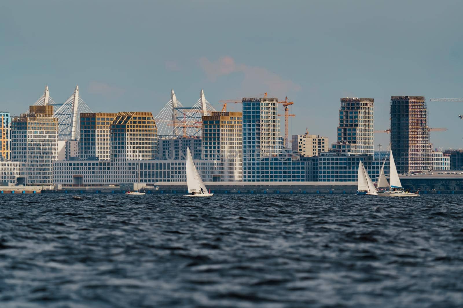 The residential complex under construction on the embankment of the Neva River on Vasilievsky island in sunny day, bulk island, construction cranes, sailing yachts, river navigation. High quality photo