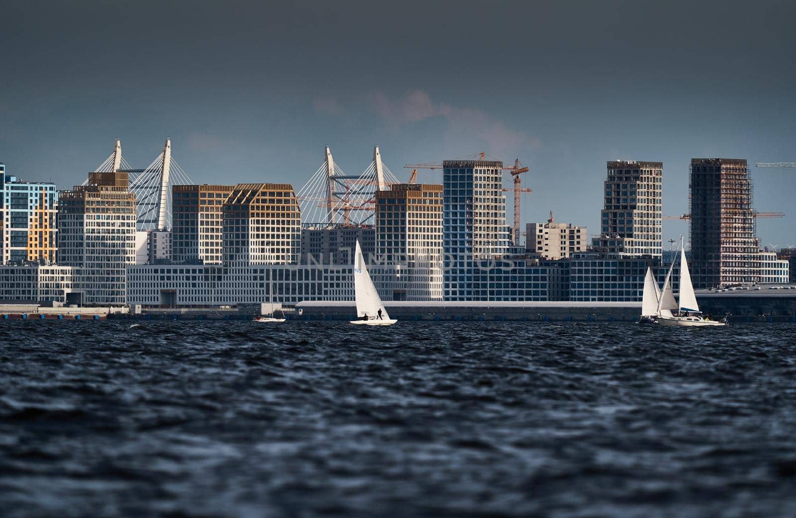 The residential complex under construction on the embankment of the Neva River on Vasilievsky island in sunny day, bulk island, construction cranes, sailing yachts, river navigation. High quality photo