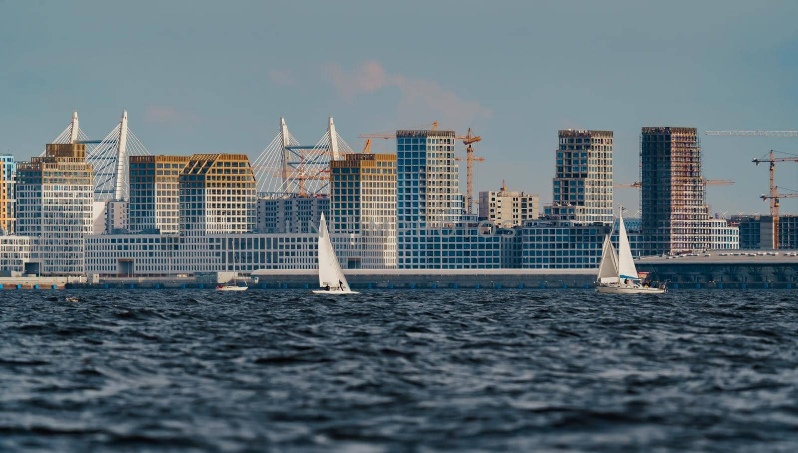 The residential complex under construction on the embankment of the Neva River on Vasilievsky island in sunny day, bulk island, construction cranes, sailing yachts, river navigation. High quality photo
