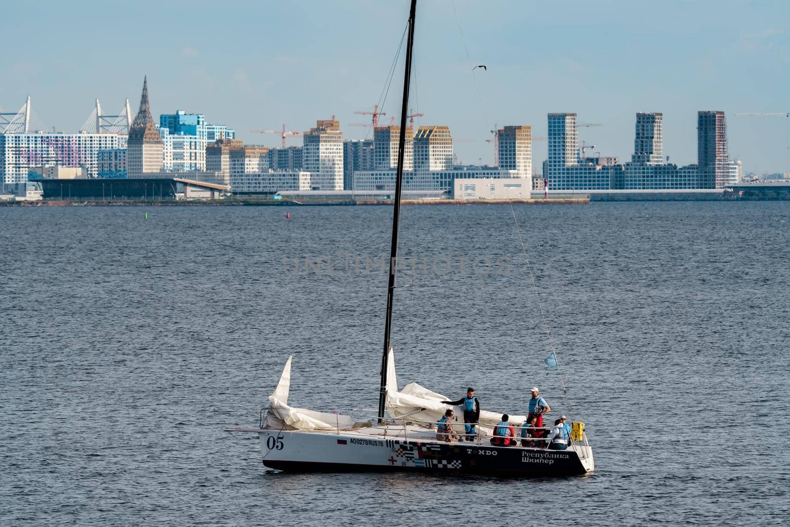 Russia, St. Petersburg, 10 June 2022: The sailing yacht with yachtsmen on board floating at sunset, residential complex under construction on the embankment on Vasilievsky island on background. High quality photo