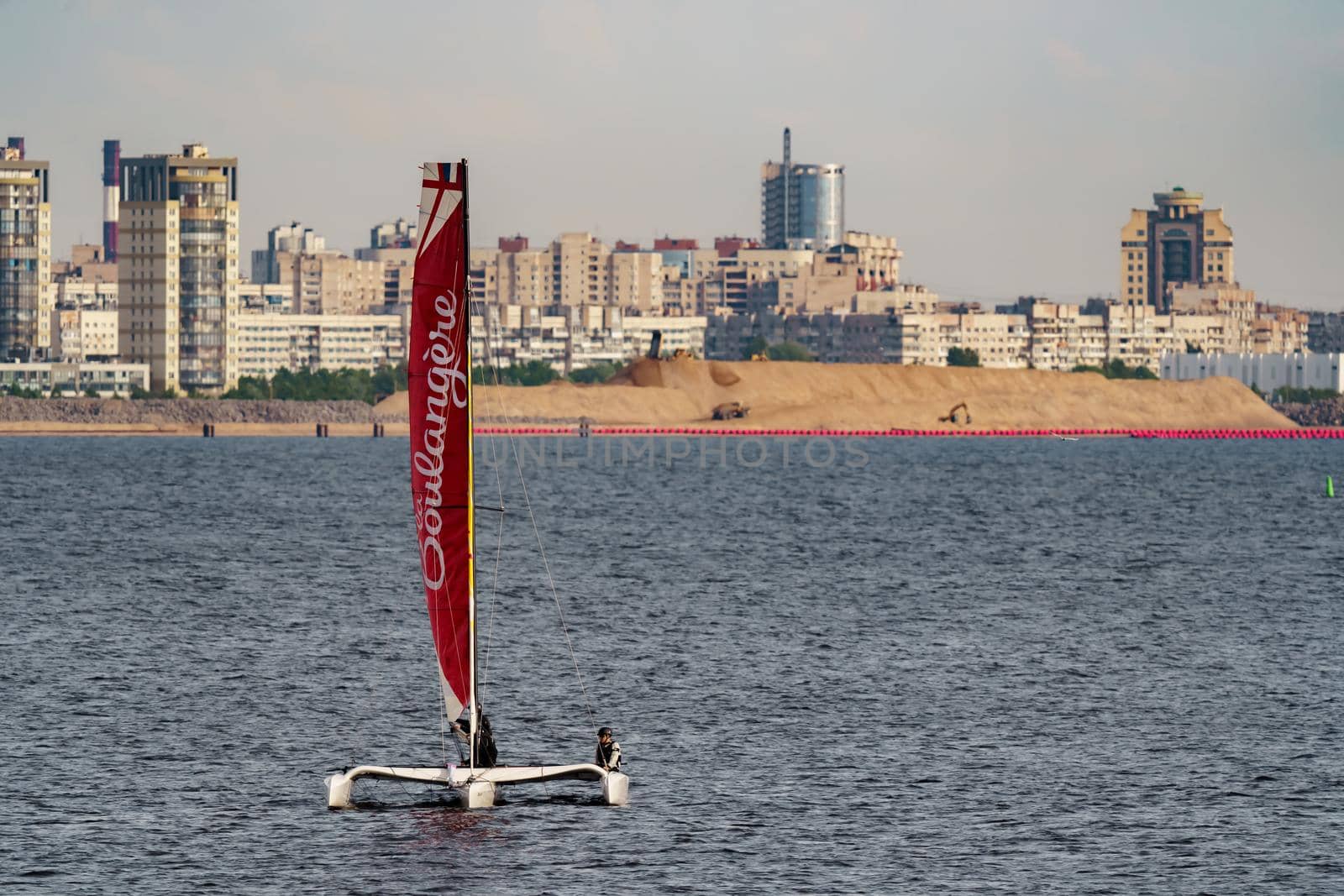 Russia, St. Petersburg, 10 June 2022: The trimaran with professional yachtsmen on board floating at sunset, residential complex under construction on the embankment on Vasilievsky island on background. High quality photo
