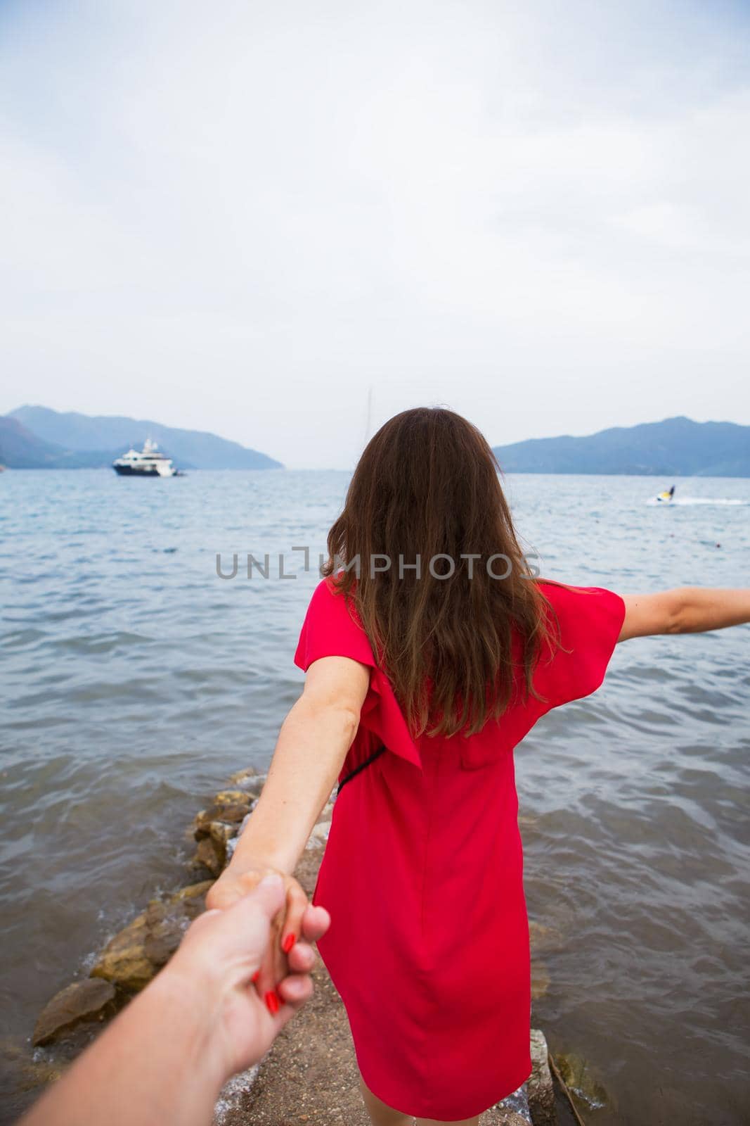 beautiful brunette girl in red dress is standing on the sea and is holding her beloved's hand by sfinks