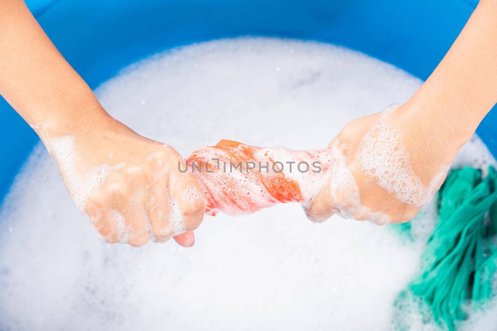 Closeup young Asian woman use hands washing color clothes in basin. Female squeeze wring out wet fabric cloth with detergent have soapy bubble in water, studio shot background, laundry concept