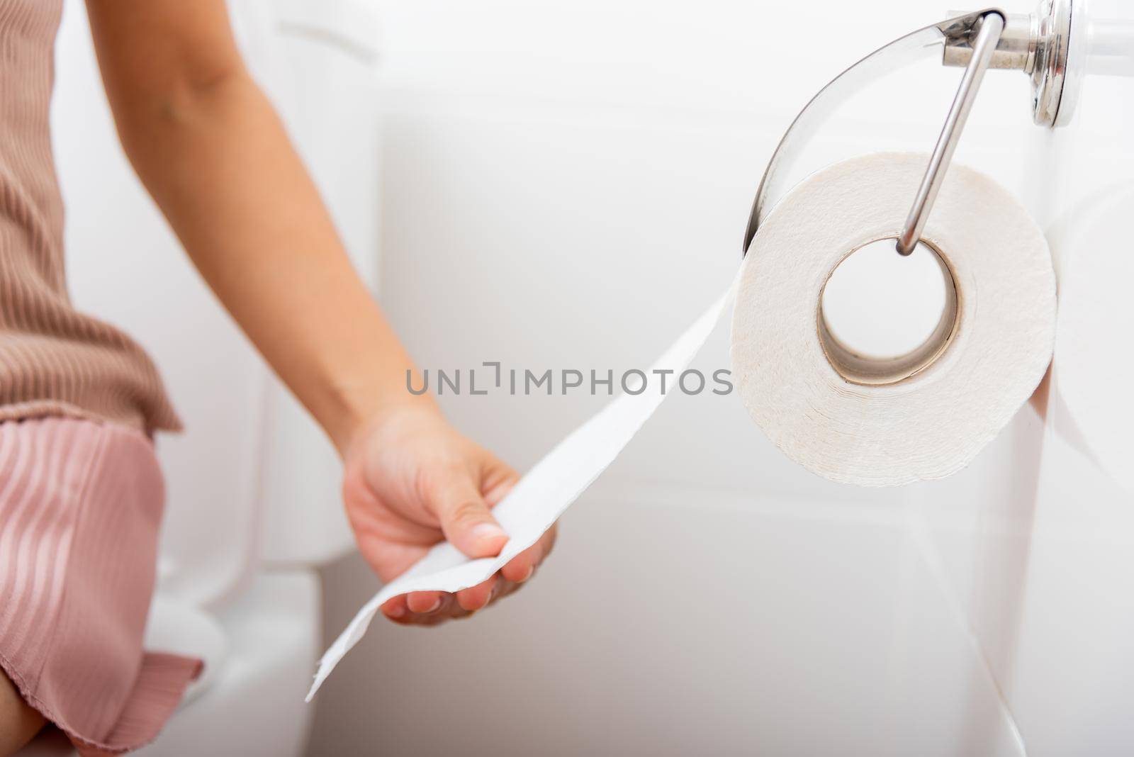 Closeup hand pulling toilet paper roll in holder for wipe, woman sitting on toilet she taking and tearing white tissue on wall to towel clean in bathroom, Healthcare concept