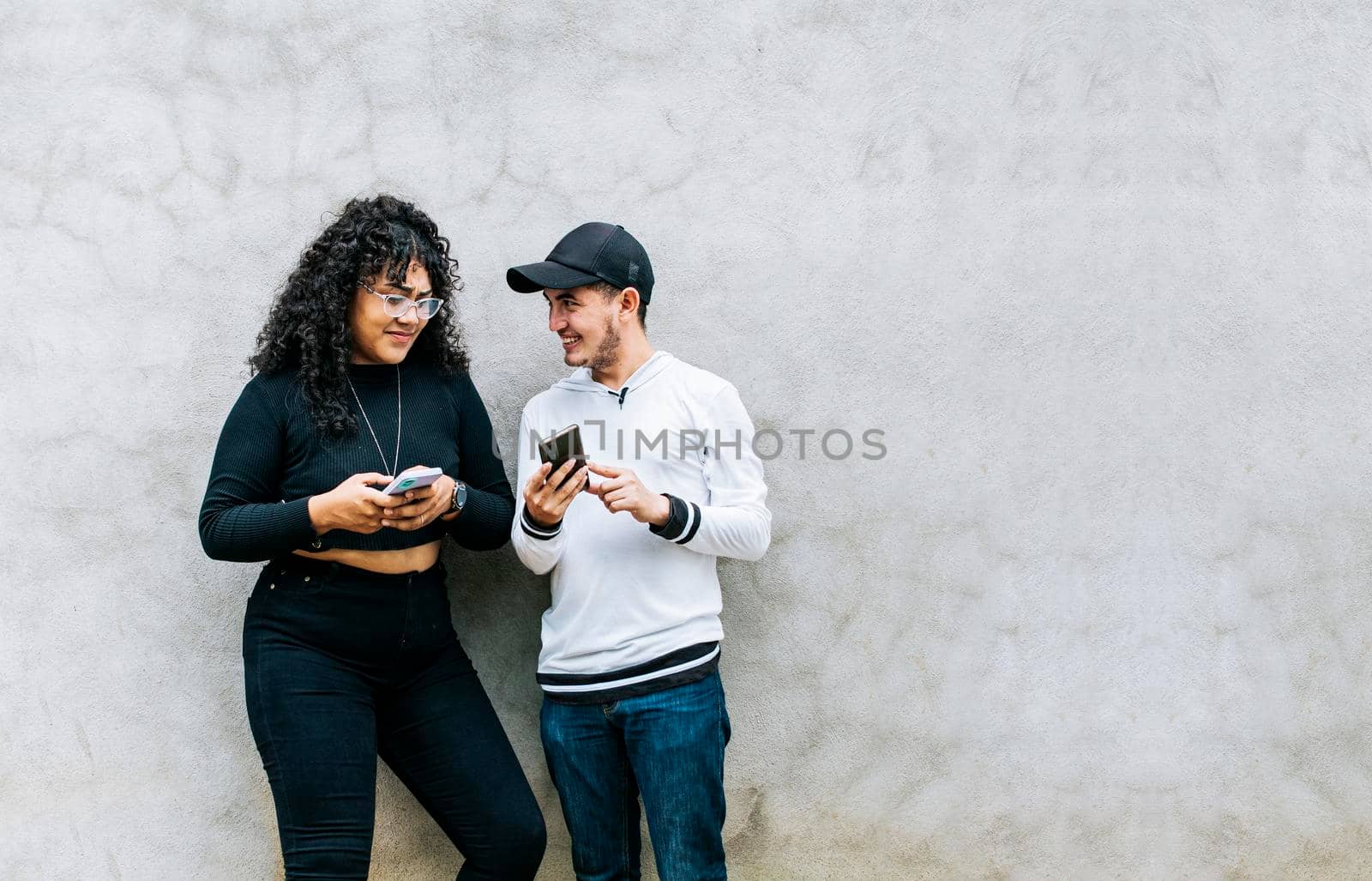 Two smiling friends checking their cell phones, Two teenage friends checking their cell phones and smiling, Two teenagers together checking their cell phones, Guy and girl leaning on a wall checking their cell phones