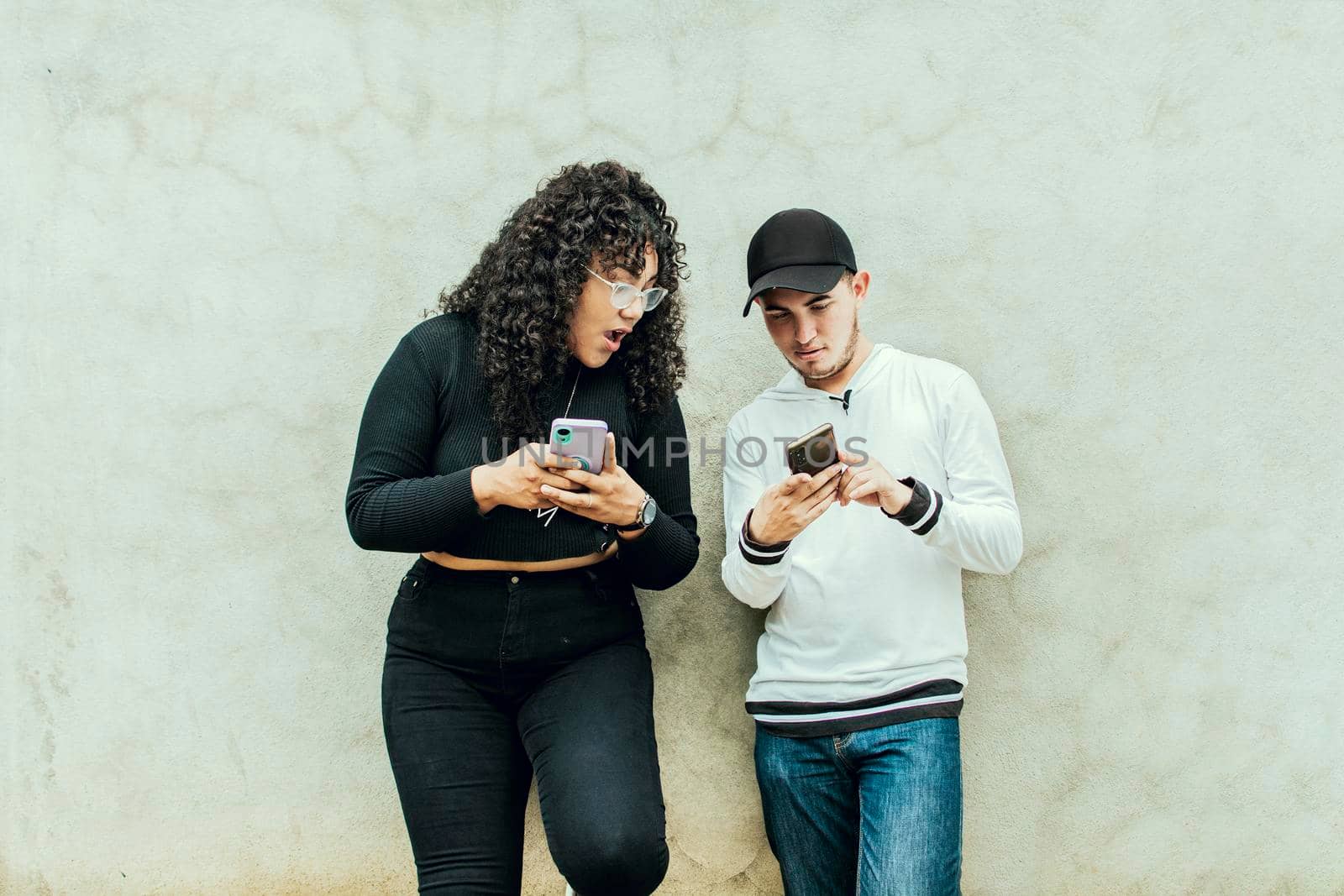 Two teenagers together checking their cell phones, Two smiling friends checking their cell phones, Two teenage friends checking their cell phones and smiling, Guy and girl leaning on a wall checking their cell phones