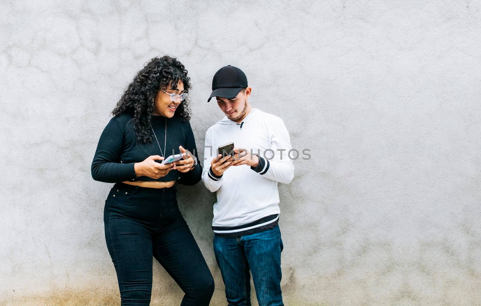 Two teenage friends checking their cell phones and smiling, Two teenagers together checking their cell phones, Guy and girl leaning on a wall checking their cell phones, Two smiling friends checking their cell phones
