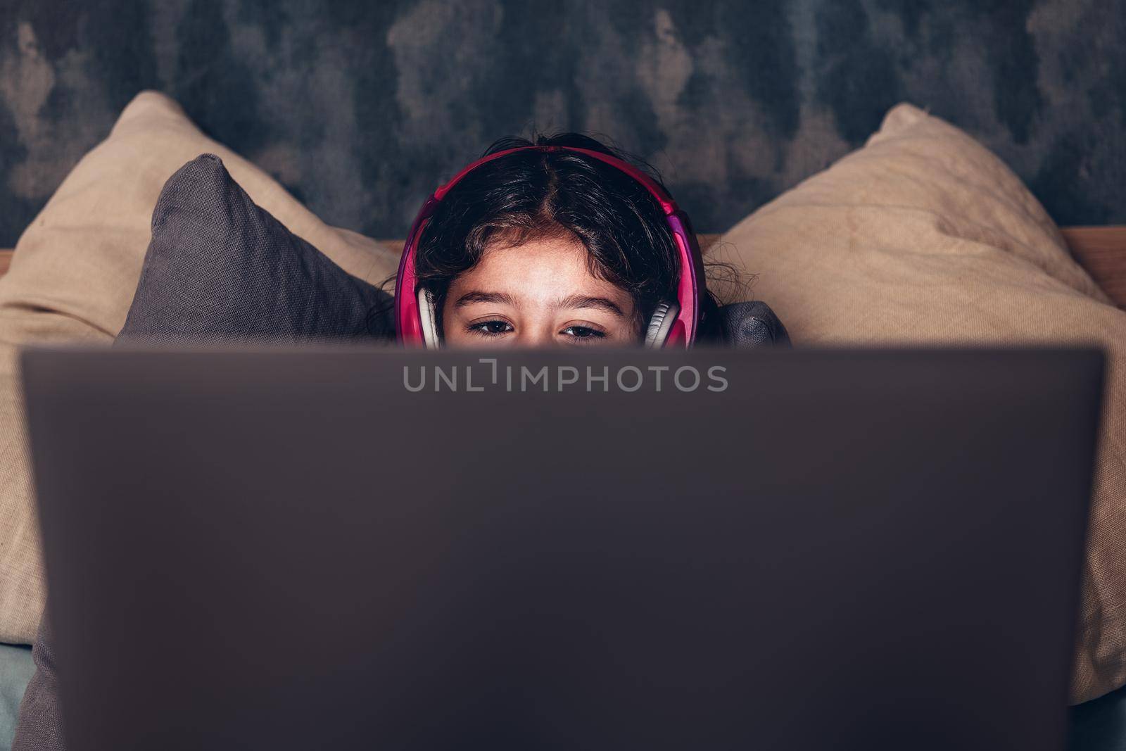 detail of the eyes of a little girl on the bed focused while playing or watching a movie on a computer in the darkness, has a pink headset, child and technology concept, copy space for text