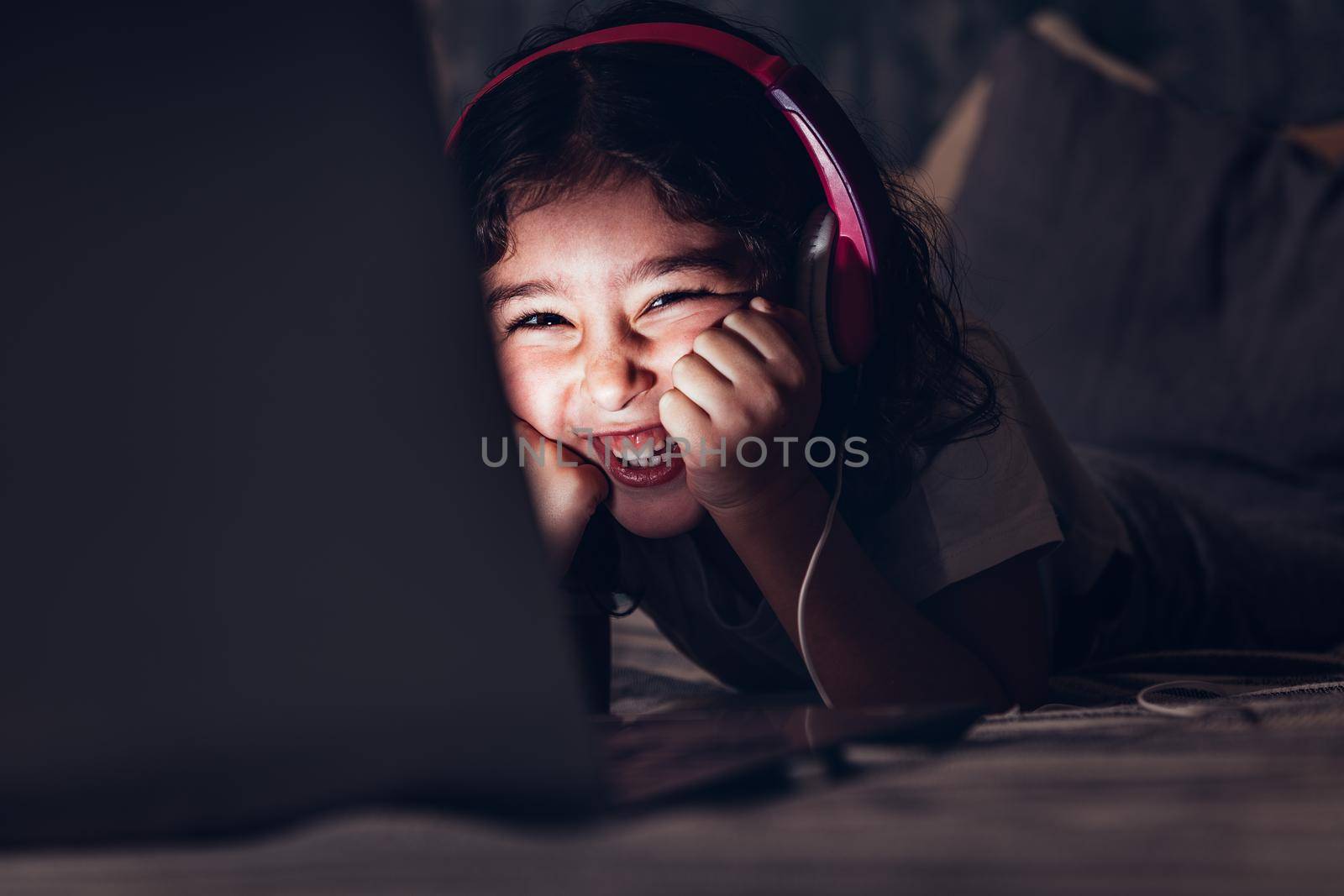 little girl lying in bed laughing while watching a movie on a laptop in the darkness, has a pink headset, child and technology concept, copy space for text