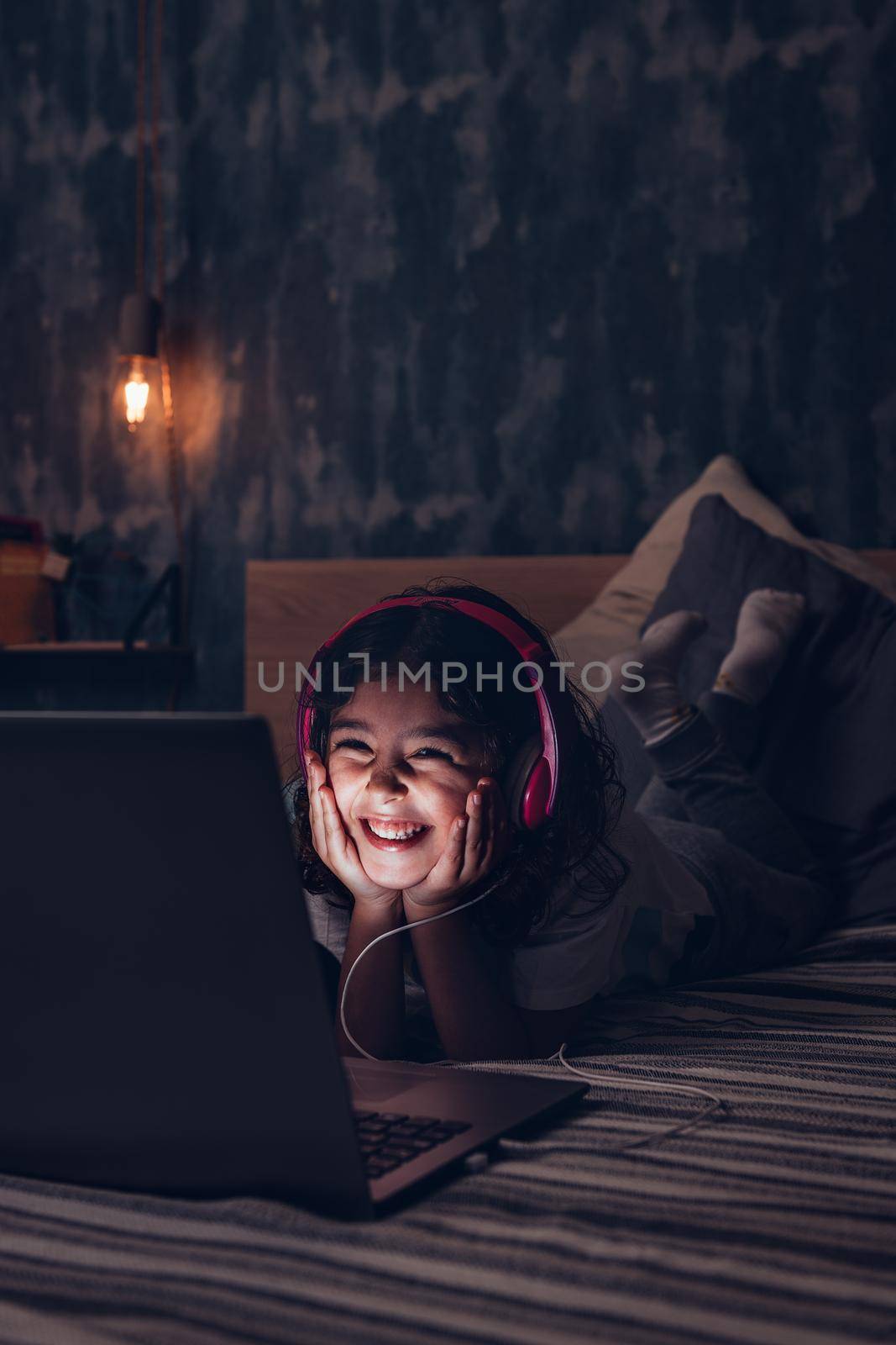 vertical photo of a happy little girl lying in bed smiling while watching a movie on a computer in the darkness, has a pink headset, child and technology concept, copy space for text
